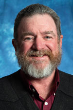 Image of Dr. Frank Hodnett, Professor in the Department of Agricultural and Extension Education at NMSU. He has brown and grey hair with a beard, and is wearing a black suit jacket over a crimson button up shirt while standing in front of a blue background.