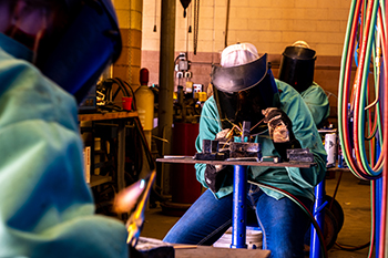 Image of a student welding in the agricultural education shop class. Student is wearing a face shield, teal protective coat, and heavy welding gloves