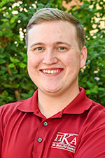 Taylor Scott headshot. He is wearing a red shirt with white writing on the right front chest. He is in front of green foliage.